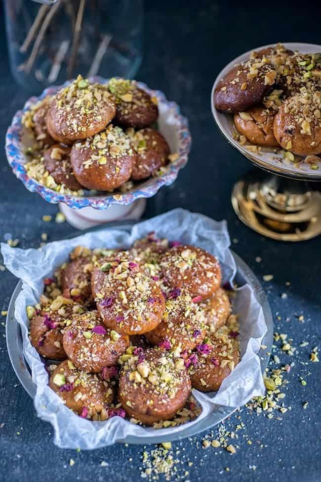 Three bowls filled with round, golden-brown Melomakarona, traditional Greek Christmas Honey Cookies topped with chopped pistachios and nuts, arranged on a dark surface. The cookies appear rich, glistening, and inviting.