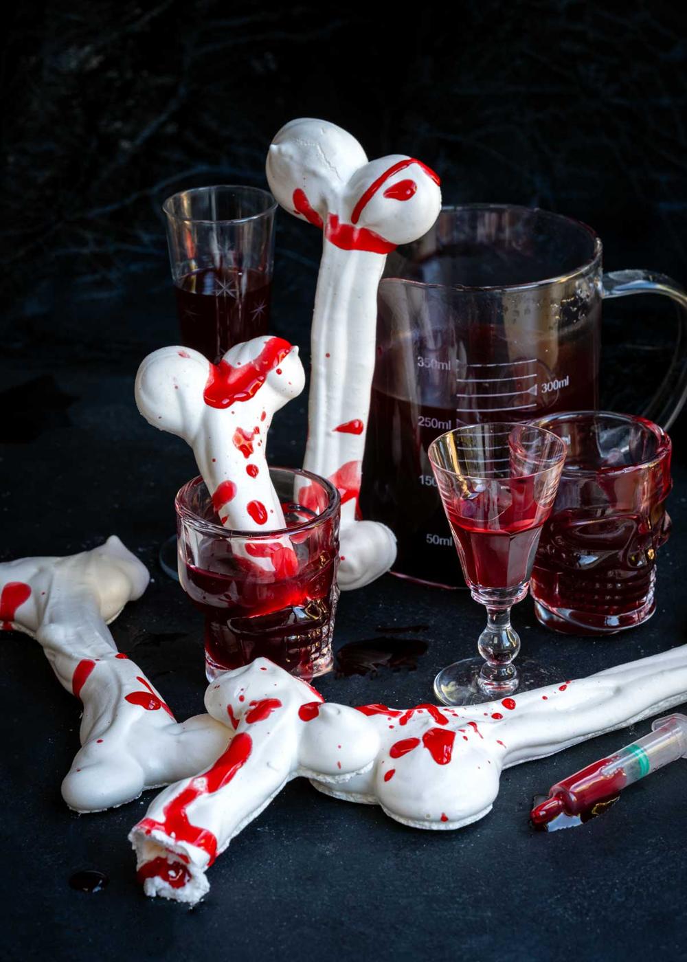 Meringue bones splattered with edible fake blood, served alongside glasses filled with dark red liquid and a plastic syringe.