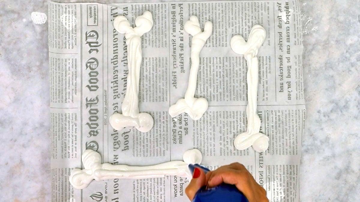 A hand piping white meringue bones with a blue piping bag onto a parchment-lined baking sheet.