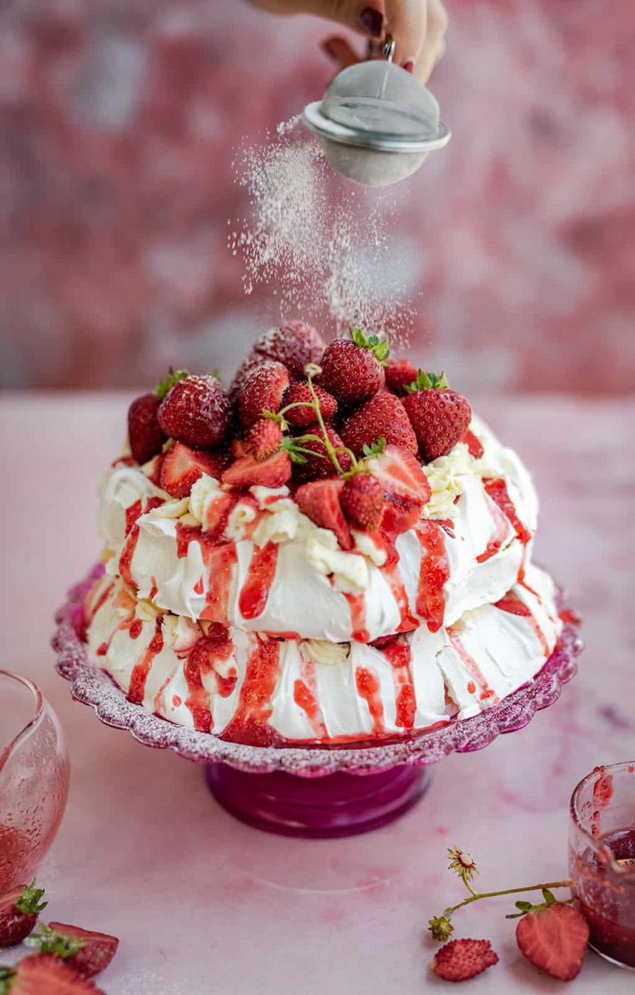 A hand sprinkles powdered sugar over a two-layer meringue cake pavlova topped with fresh strawberries and strawberry sauce, displayed on a pink glass cake stand against a pink background.