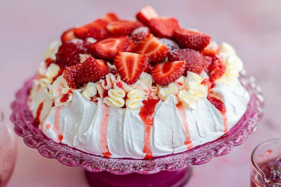 A meringue cake pavlova topped with whipped cream and fresh strawberry slices, drizzled with strawberry sauce, sits on a pink glass cake stand. Scattered strawberries and a jar of sauce are nearby on a pink surface.