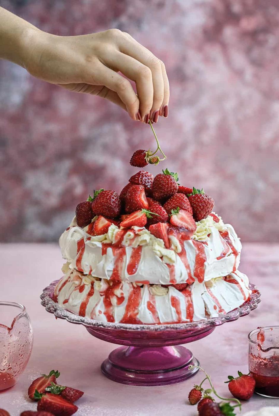 A hand places a fresh strawberry atop a two-layer meringue cake covered with whipped cream, strawberry sauce, and whole strawberries, beautifully displayed on a pink glass cake stand against a pink background.