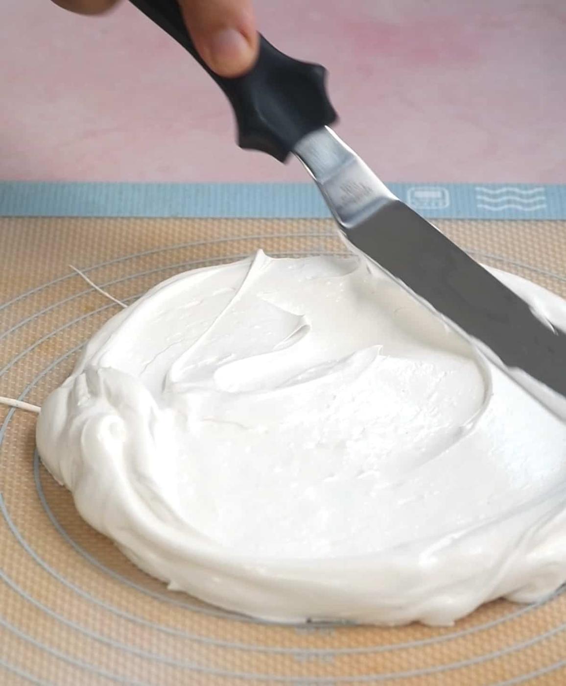 A hand uses an offset spatula to spread fluffy white meringue for a meringue cake onto a silicone baking mat laid on a wire cooling rack.