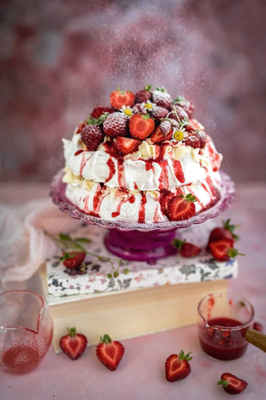 A two-layer meringue cake topped with whipped cream, fresh strawberries, and a drizzle of red sauce, displayed on a pink cake stand. Strawberry slices and sauce are scattered around on a pink surface.