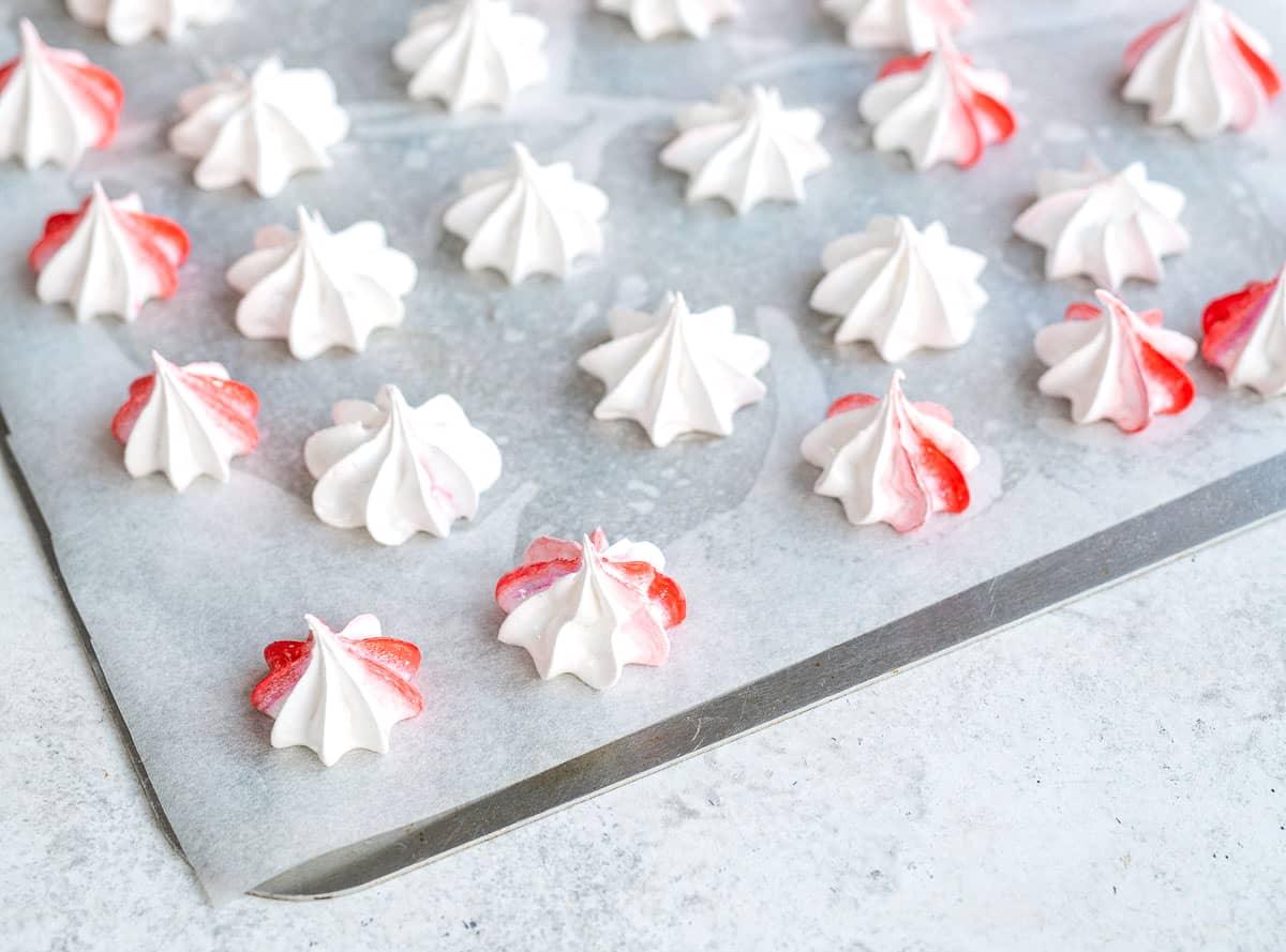 Rows of small white Meringue Kisses, some with red streaks, are piped onto a sheet of parchment paper on a baking tray. The surface underneath is a light gray countertop.