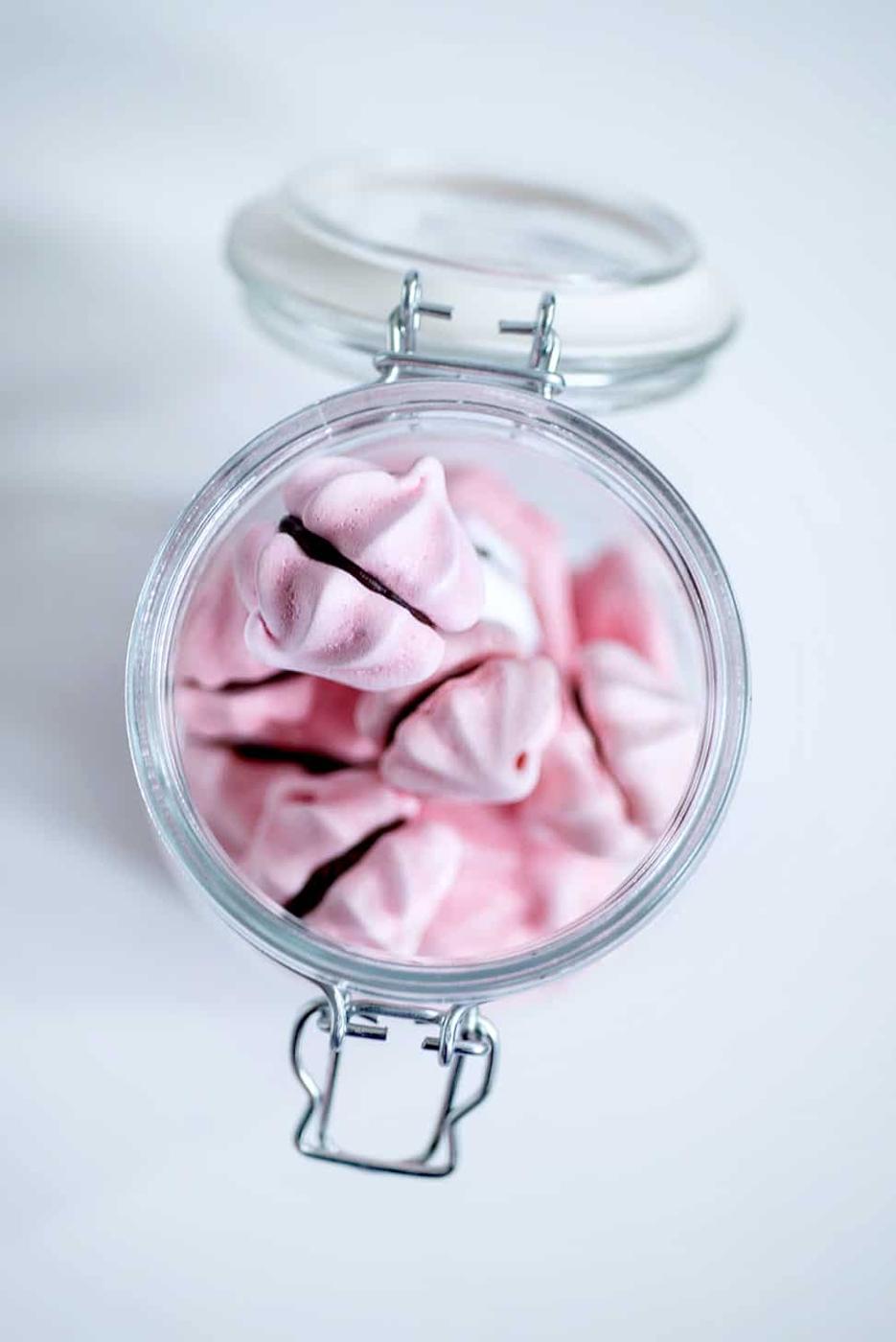 A glass jar filled with delicate meringue cookies, viewed from above. The open lid reveals cookies neatly arranged inside, their pink hues contrasting beautifully with the white background. Perfect inspiration for your next meringue cookie recipe.