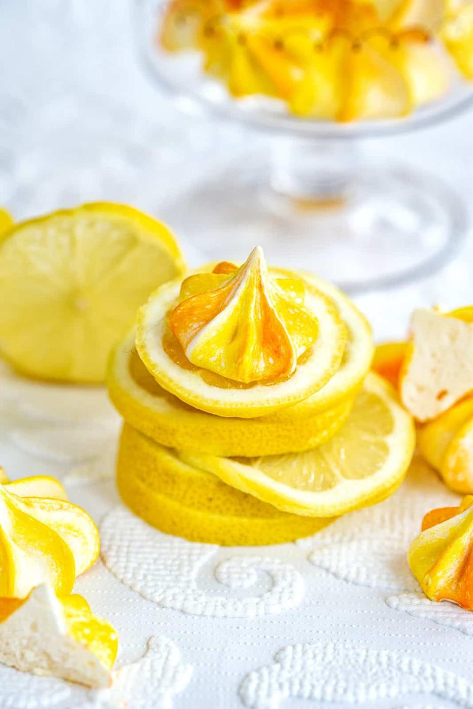 Slices of lemon stacked with a yellow and white meringue cookie on top, surrounded by more lemon slices and meringue cookies on a white textured surface. A glass dish with additional meringues is in the background.