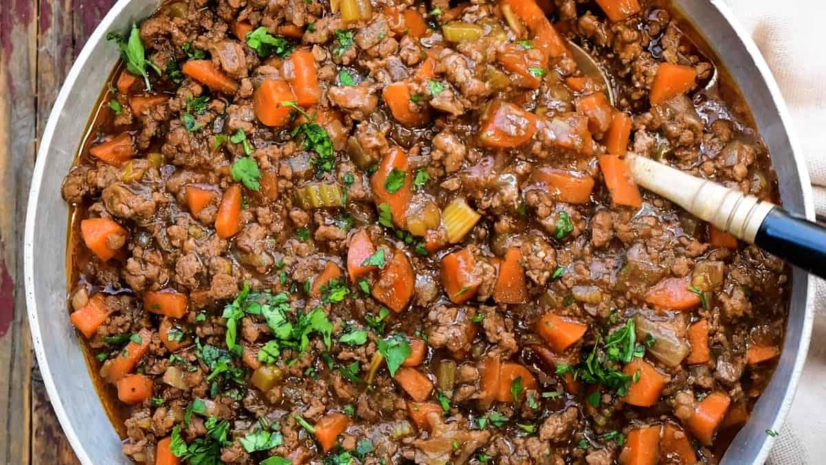A skillet filled with cooked ground beef, chopped carrots, celery, and fresh herbs in a rich brown sauce—a classic Scottish recipe reminiscent of Mince and Tatties—being stirred with a spoon.