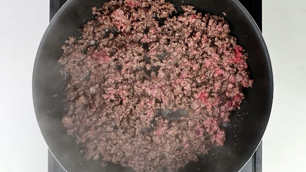 Ground beef is being browned in a black frying pan on a stovetop, with some pieces still showing a bit of pink. Steam rises from the cooking meat, perfect for starting a classic Scottish recipe like Mince and Tatties.