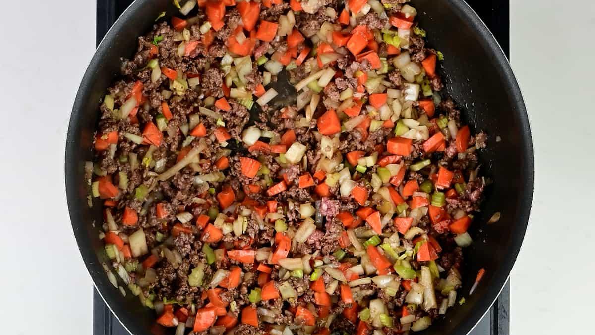 A frying pan filled with chopped onions, carrots, and ground beef is being sautéed on a stovetop. The ingredients are mixed and partially cooked, creating the savory base for a classic Scottish recipe like Mince and Tatties.