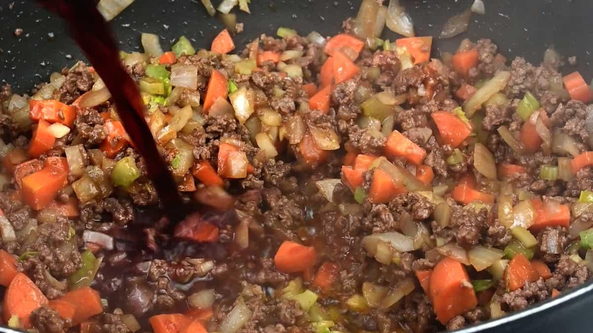 Ground beef, chopped carrots, celery, and onions simmer in a pan as red wine is poured in, creating a rich and savory mixture—perfect for a classic Scottish recipe like Mince and Tatties.
