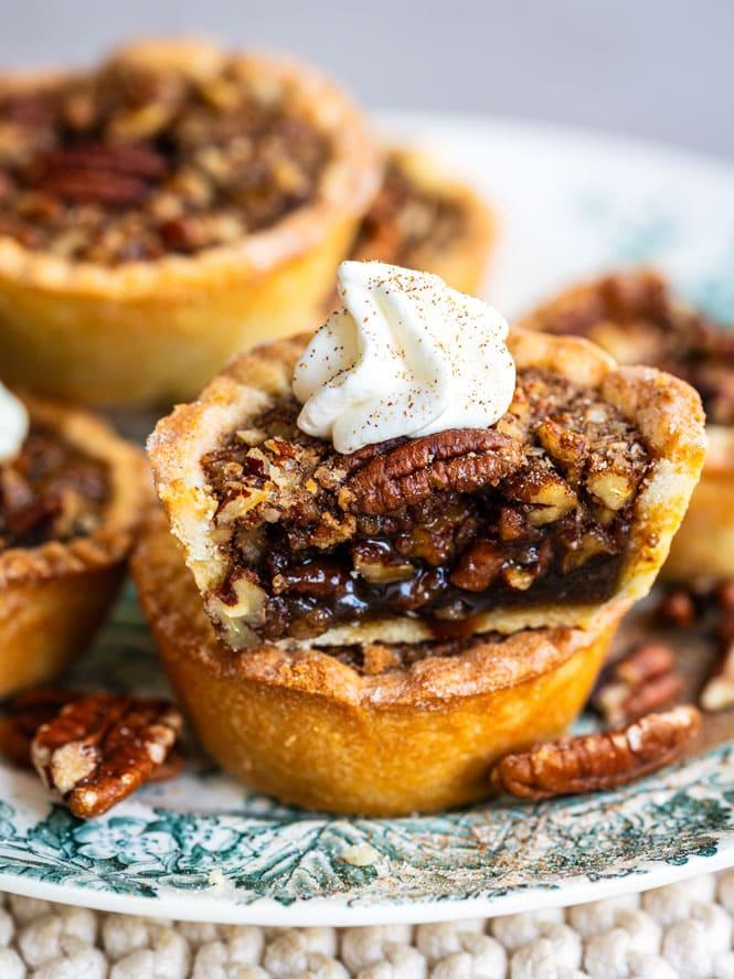 A close-up of mini pecan pies, one cut in half to show the gooey filling and topped with a dollop of whipped cream and a pecan. Other pecan pies and pieces of pecan are visible in the background.