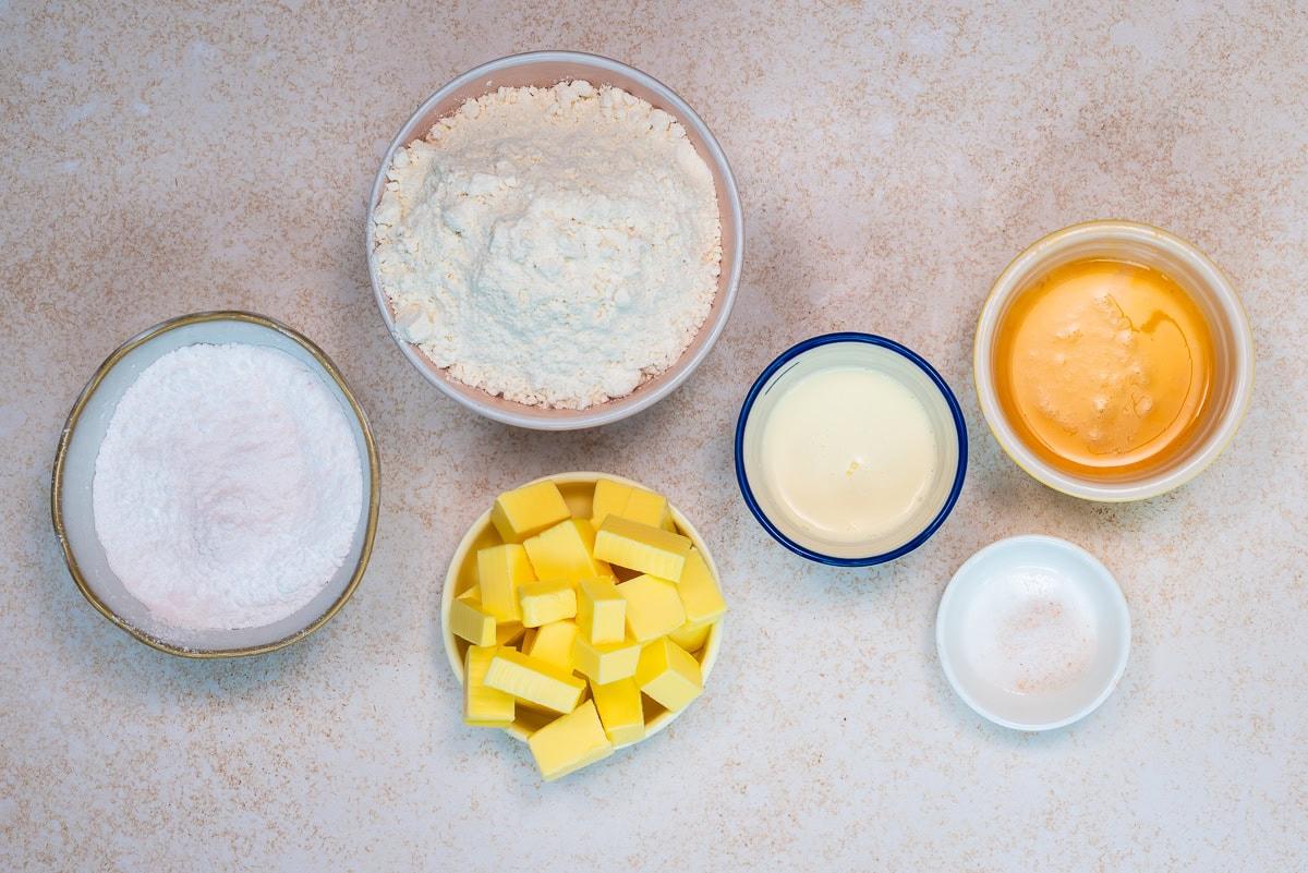 Six bowls on a light surface containing baking ingredients: flour, powdered sugar, cubed butter, sweetened condensed milk, an egg, and a small amount of salt.