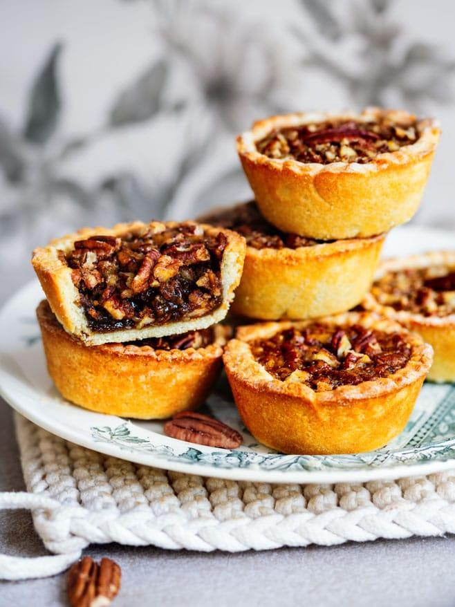 A plate of six pecan butter tarts, with one tart cut in half revealing a gooey nut filling. The tarts sit on a patterned plate atop a textured cloth, with pecans scattered nearby.