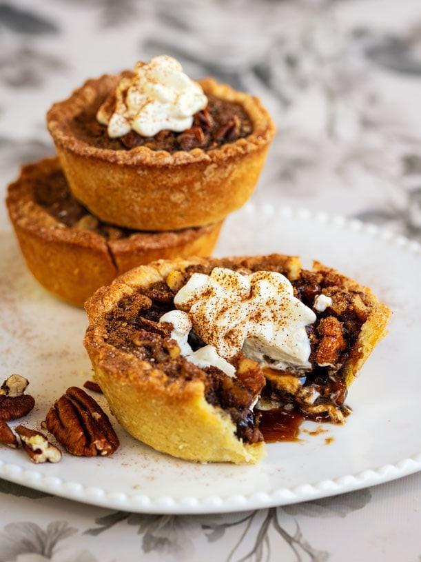 Two mini pecan pies, one cut open, sit on a white plate. Both are topped with whipped cream and a sprinkle of cinnamon. Chopped pecans are scattered nearby. The background is softly blurred.