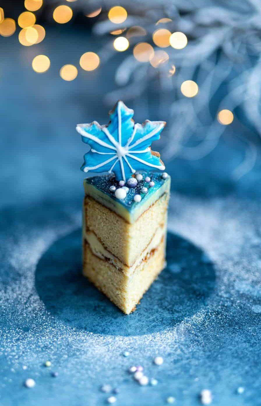 A square slice of layered Mirror Glaze Cake topped with silver sprinkles and a blue snowflake-shaped icing decoration, set against a blue background with blurred festive lights.