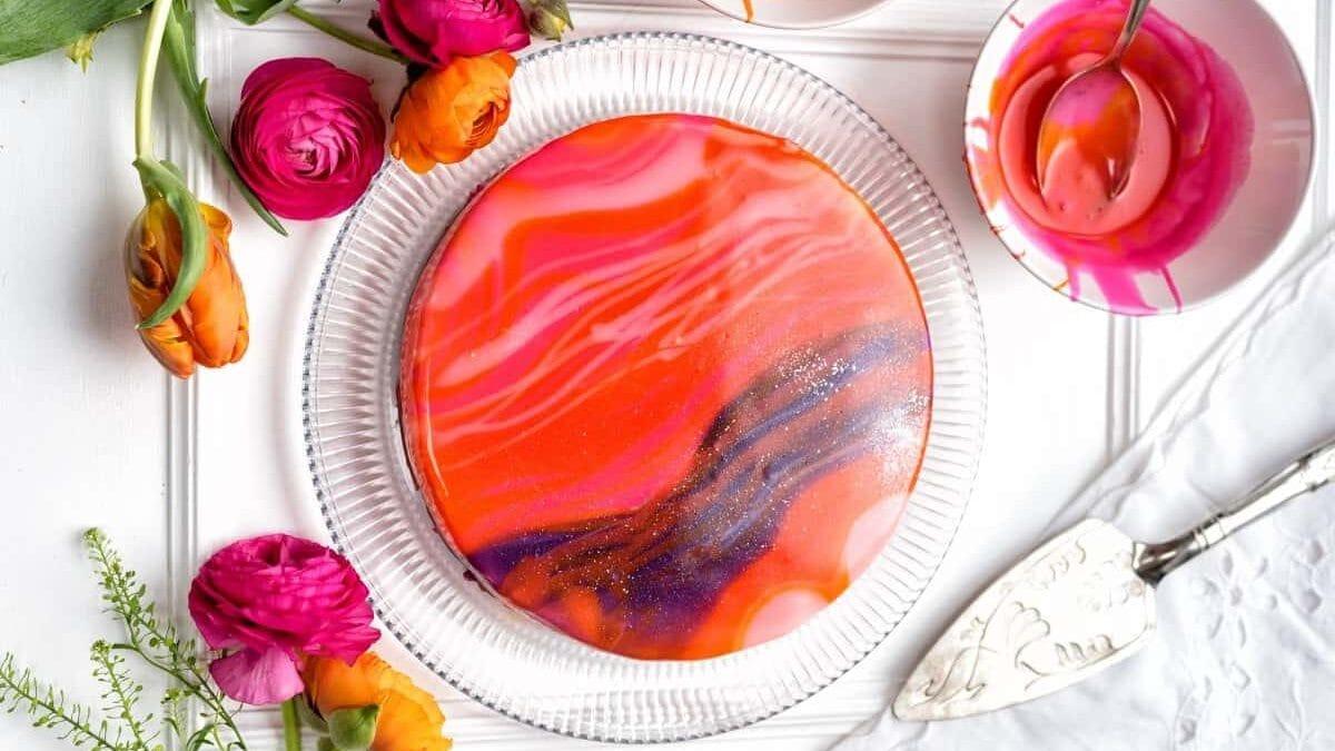 A round mirror glaze white chocolate cheesecake with a glossy, marbled finish in pink, red, orange, and purple sits on a glass plate, surrounded by colorful flowers and bowls of glaze on a white surface.