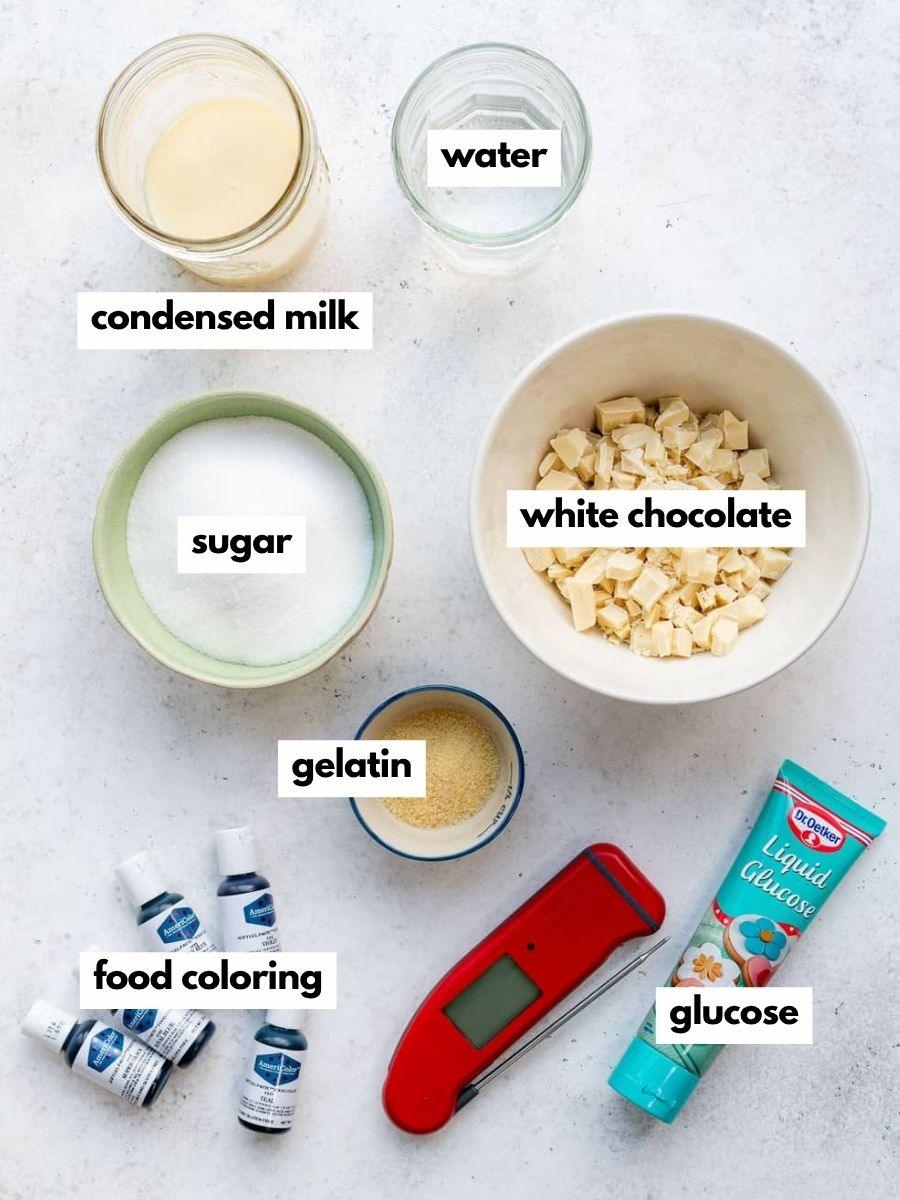 An overhead view of ingredients for a Mirror Glaze Cake recipe, including bowls of condensed milk, water, sugar, white chocolate, gelatin, bottles of food coloring, a tube of glucose, and a red digital thermometer on a white surface.