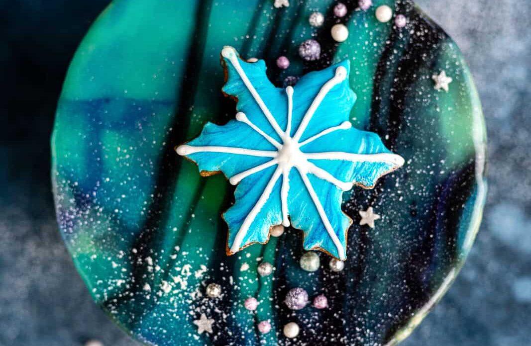 A Mirror Glaze Cake with blue, green, and black marbled frosting, topped with a large blue snowflake cookie, silver and purple edible pearls, white sprinkles, and small star-shaped decorations.