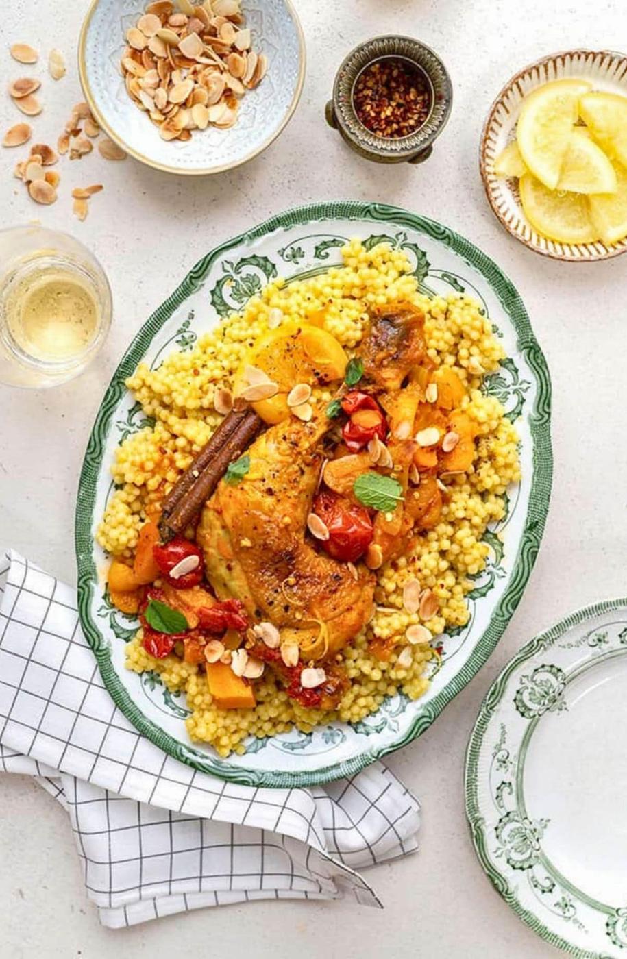 A platter of Moroccan Chicken couscous topped with braised chicken, tomatoes, squash, almonds, and herbs, surrounded by bowls of sliced almonds, lemon wedges, and seasonings on a light table with a checkered napkin.