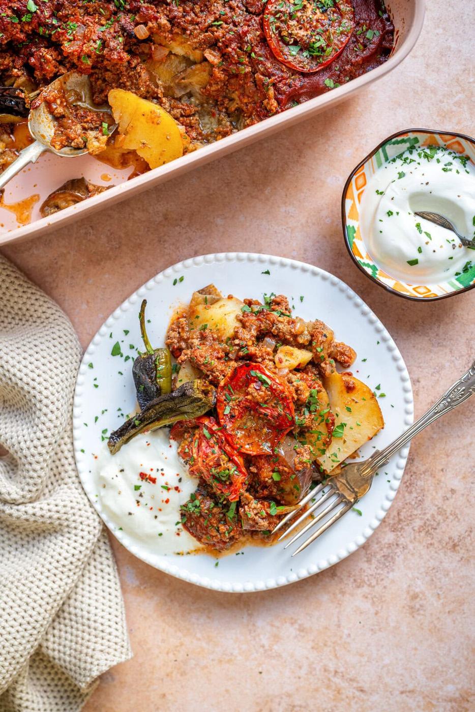 A plate of mousakka with ground meat, potatoes, tomatoes, and peppers, served with a side of creamy white yogurt. The casserole dish and a linen napkin are nearby on a light-colored surface.