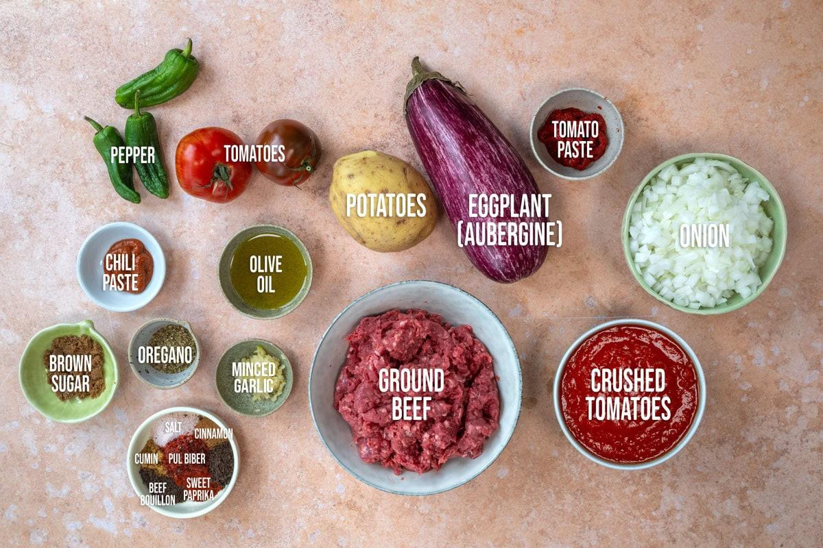 Various labeled ingredients for a Musakka recipe are arranged on a countertop, including ground beef, eggplant, potato, tomatoes, onion, spices, olive oil, brown sugar, chili paste, tomato paste, and crushed tomatoes.