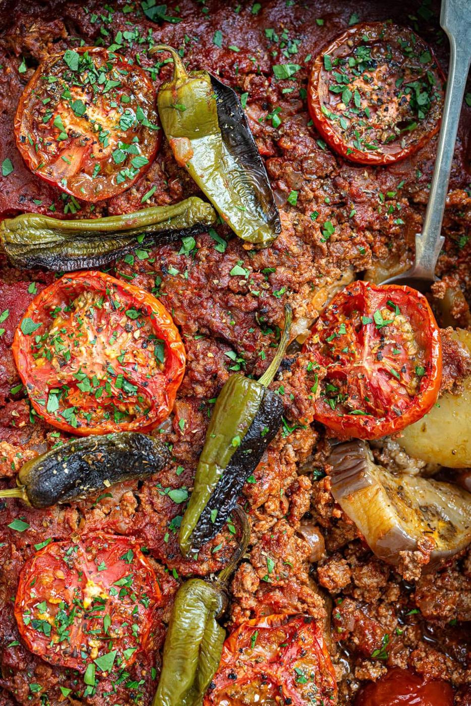 Close-up of a baked musakka dish with ground meat, sliced tomatoes, whole roasted green peppers, and chopped herbs on top, with a serving spoon partially visible on the right side.