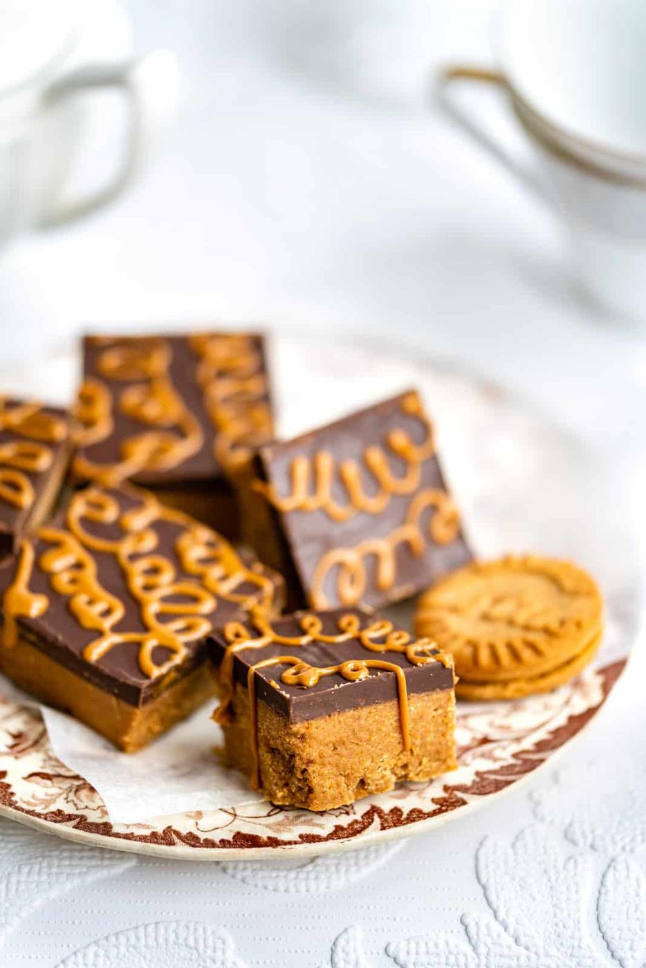 A plate with several no bake Biscoff Bars, each chocolate-topped and drizzled with caramel, sits next to a sandwich cookie. Teacups and a white background complete the inviting scene.