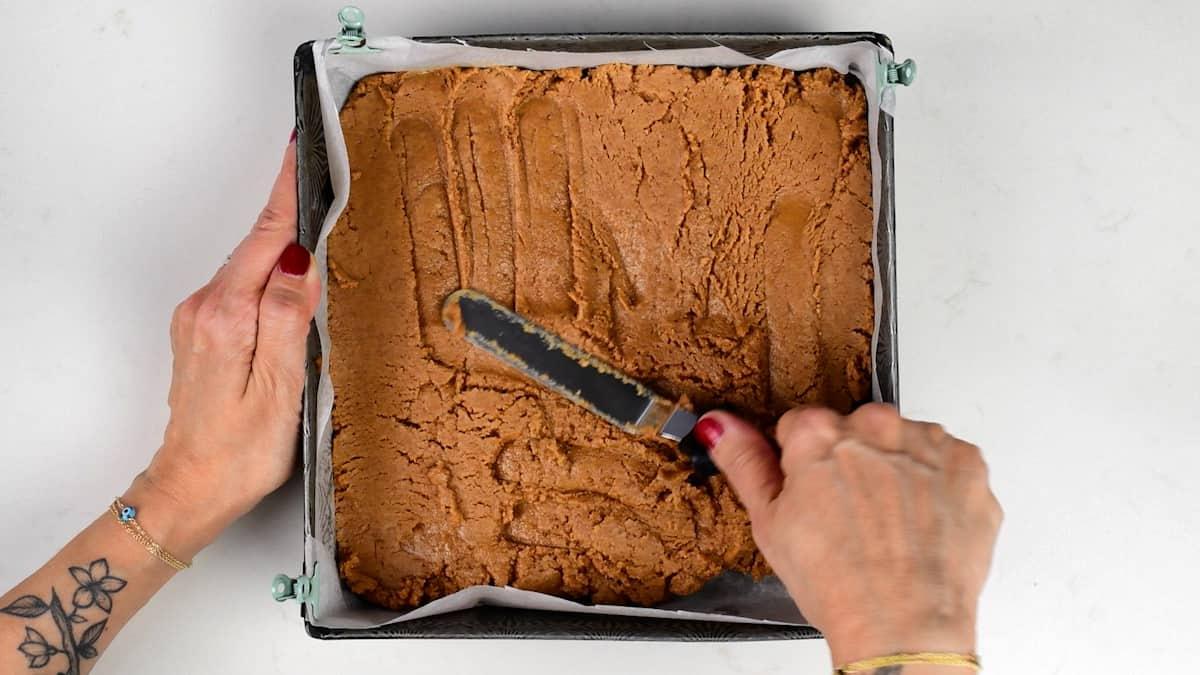 A person spreads thick brown batter for Biscoff Bars in a square baking pan lined with parchment paper using an offset spatula. Their fingernails are painted red, and one wrist has a flower tattoo—a delicious no bake treat in the making.