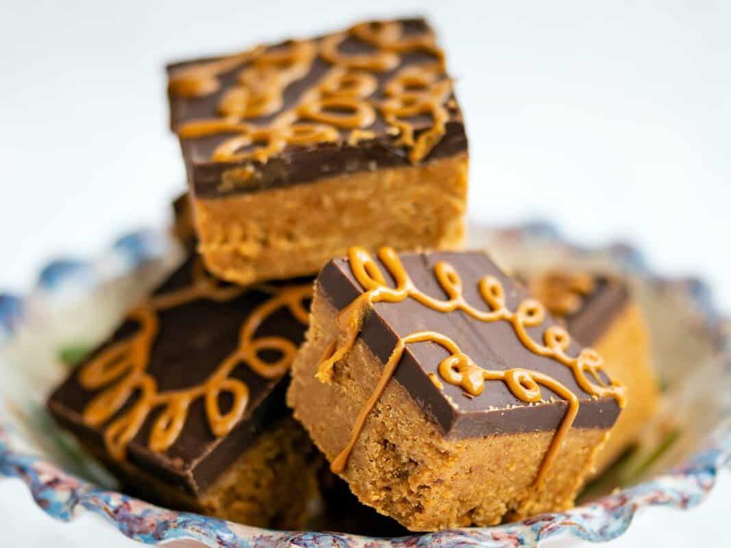 A decorative pedestal plate holds stacked no bake Biscoff Bars featuring a chocolate layer and orange drizzle, revealing a thick, crumbly base. The background is softly blurred.