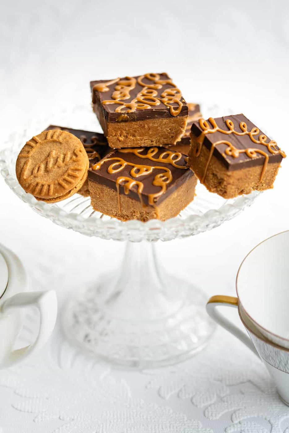 A glass cake stand holds no bake Biscoff Bars topped with chocolate and caramel drizzle, alongside a round cookie, surrounded by two white teacups with gold rims on a white surface.