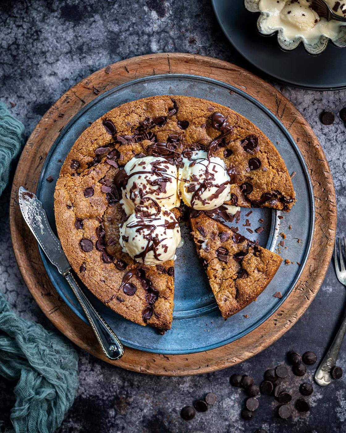 A giant chocolate chip cookie topped with three scoops of vanilla ice cream and chocolate drizzle sits on a plate. One slice is cut and slightly pulled out; a knife and chocolate chips are nearby.