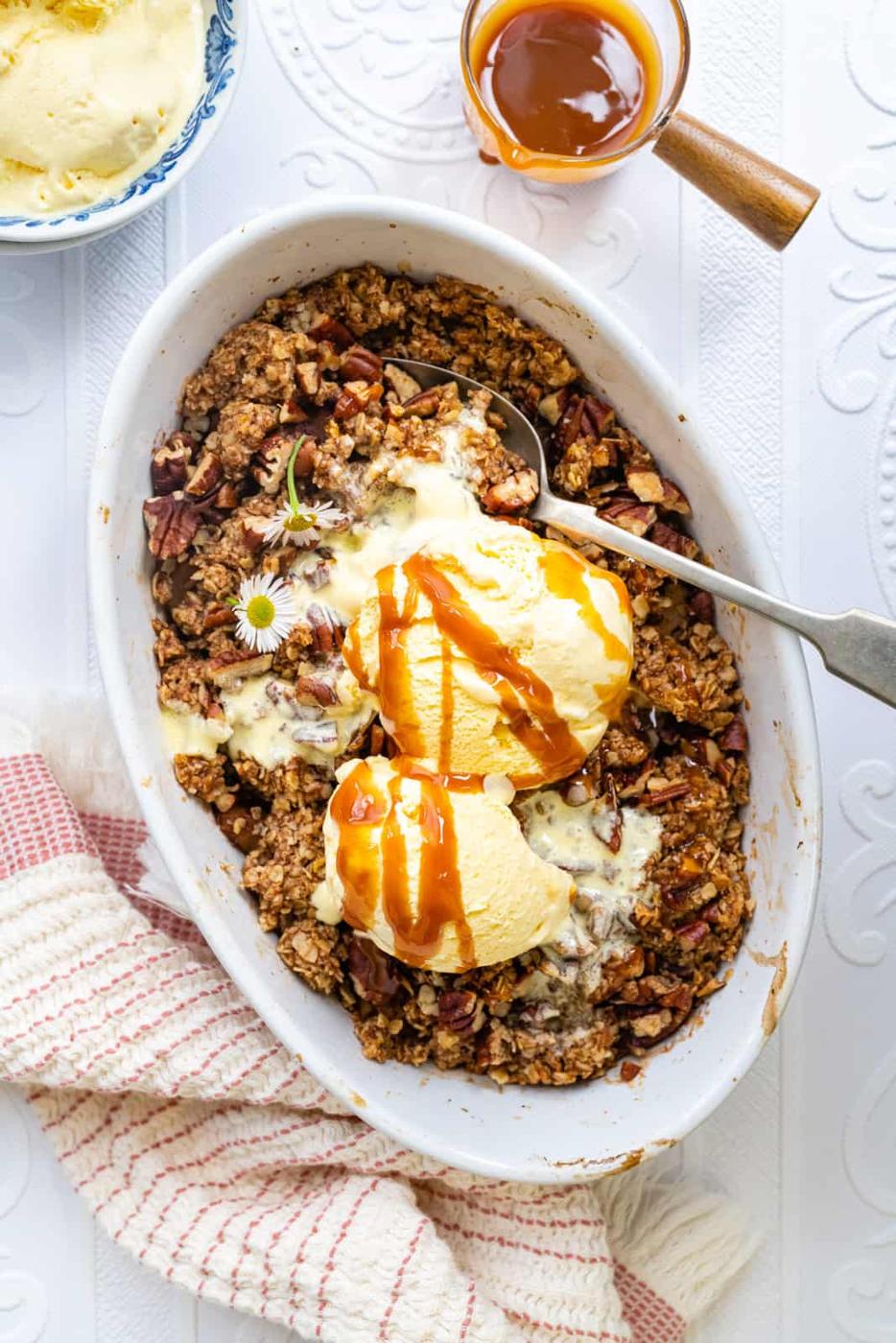 An oval white dish holds an Apple Crumble dessert with a nutty oat topping, topped with two scoops of vanilla ice cream and caramel sauce. A spoon and a cup of caramel are nearby, with a red-striped cloth on the table.