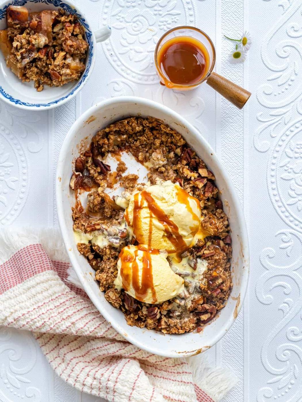 Overhead view of an apple crumble dish with oat topping, two scoops of vanilla ice cream, and caramel sauce, alongside a small bowl of apple crisp, a cup of caramel sauce, and a striped towel on a white textured surface.