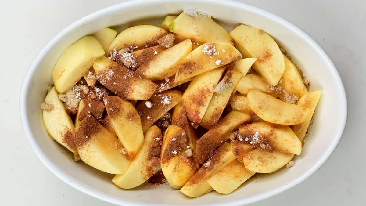Sliced apples arranged in a white oval baking dish, topped with brown sugar, cinnamon, and a classic oat topping for a delicious apple crumble, ready to be baked.