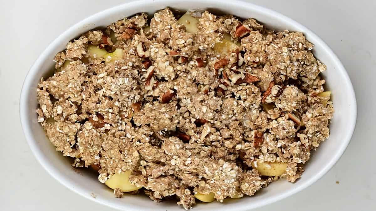 An oval white dish filled with an unbaked apple crumble is topped with a crumbly oat topping. The dessert sits on a plain white surface, ready to be baked to golden perfection.