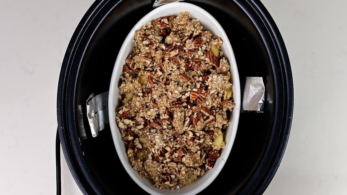 An oval white dish filled with Apple Crumble and a crumbly oat topping sits inside a black slow cooker, ready to be cooked. The slow cooker is viewed from above on a white background.
