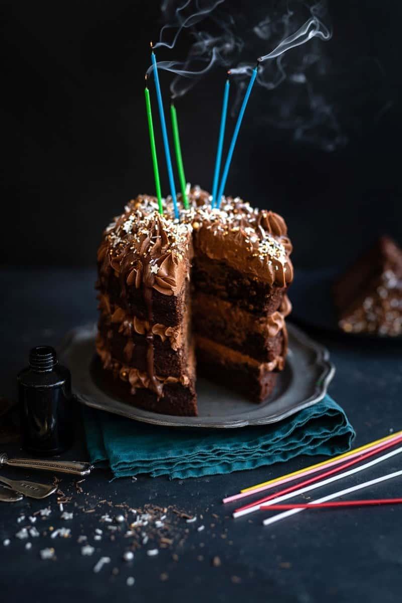 A decadent Black Magic Cake with chocolate frosting and sprinkles sits on a plate. Four colorful candles, just blown out, send wisps of smoke skyward. In the background, a slice of cake rests on a dark surface.