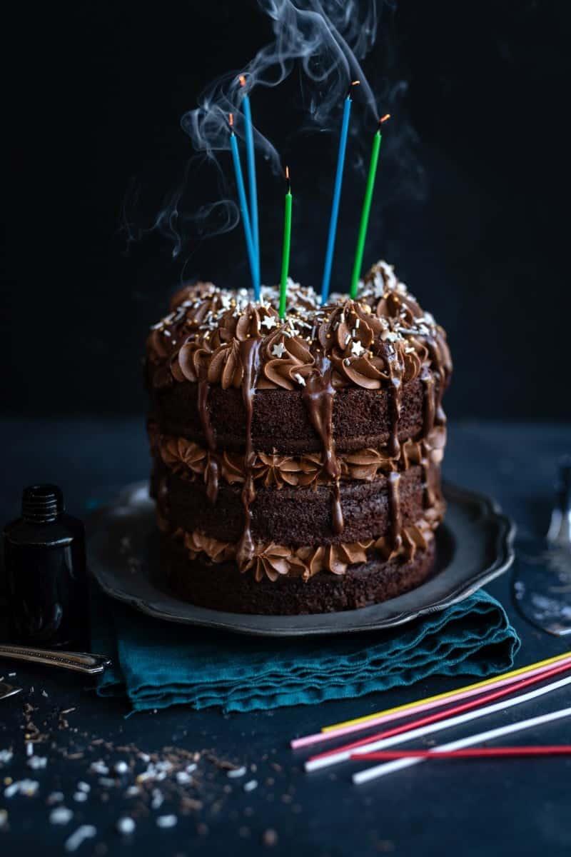 A decadent Black Magic Cake, layered with chocolate frosting and sprinkles, is topped with five smoking birthday candles. The cake sits on a plate against a dark background, with unlit candles scattered nearby.