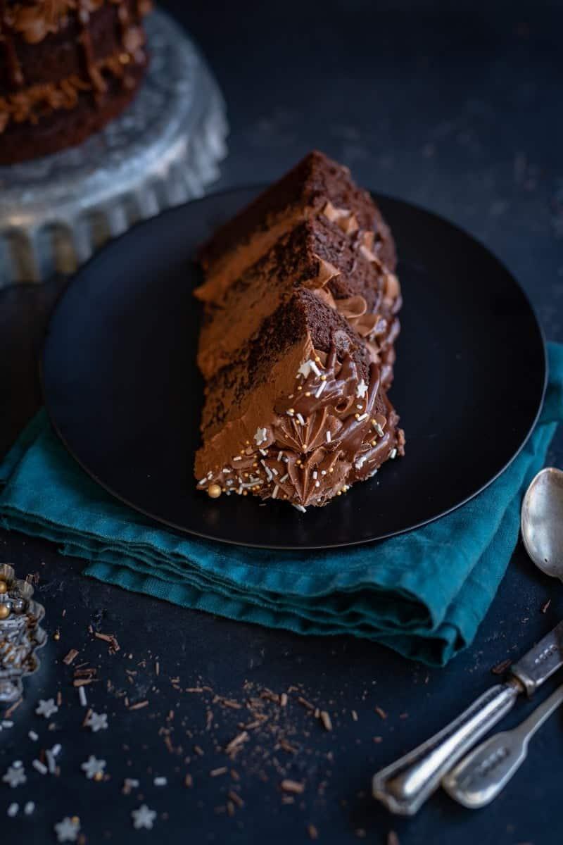 A slice of rich Black Magic Cake with chocolate frosting and sprinkles sits on a black plate, placed on a dark teal napkin, with a fork and spoon nearby on a dark surface.