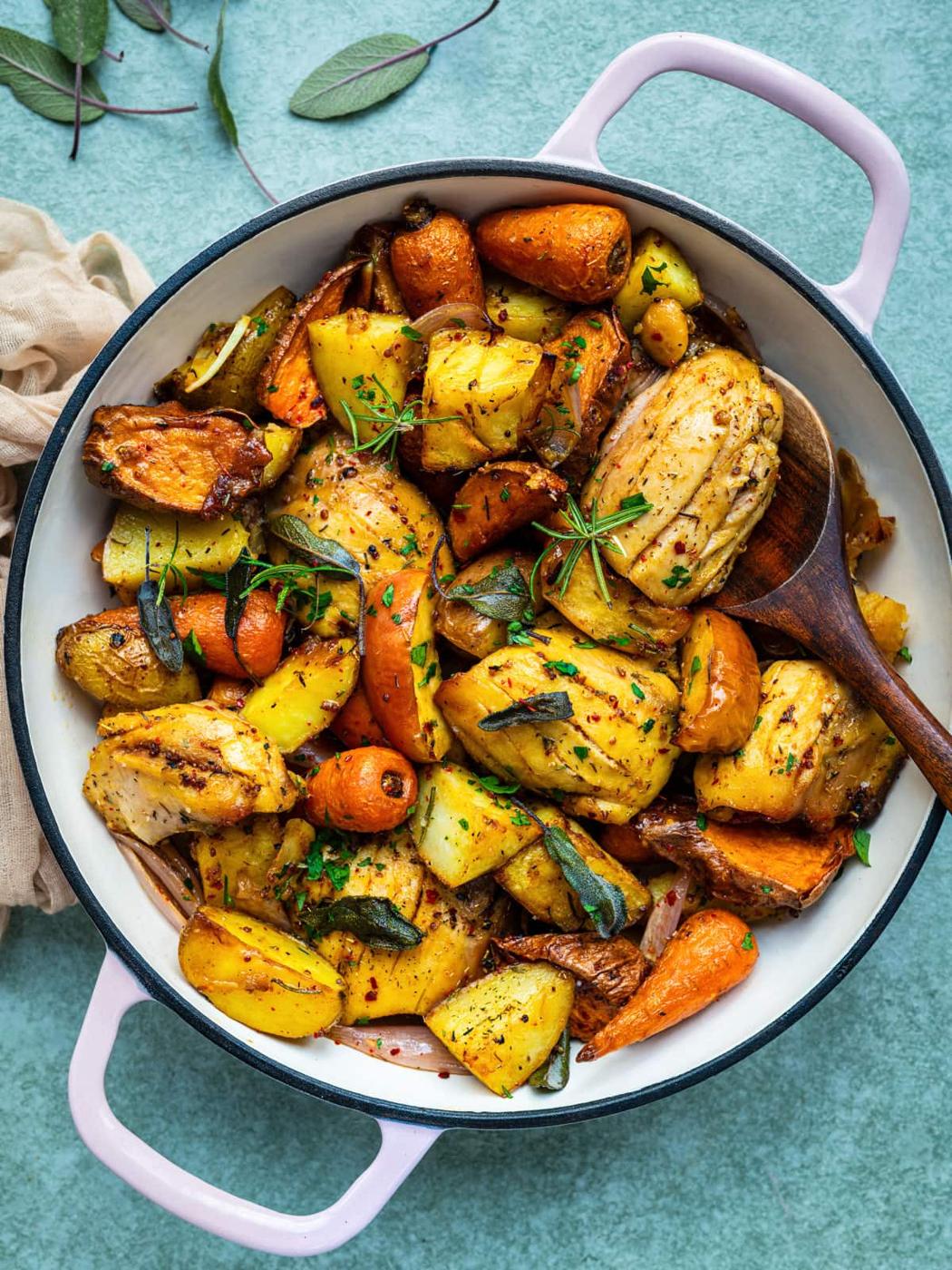 A round white pot with pink handles filled with roasted vegetables, including potatoes, carrots, and herbs, sits on a light green surface. This one pan autumn chicken dinner can be made easily in an air fryer or oven. A wooden spoon rests in the pot.