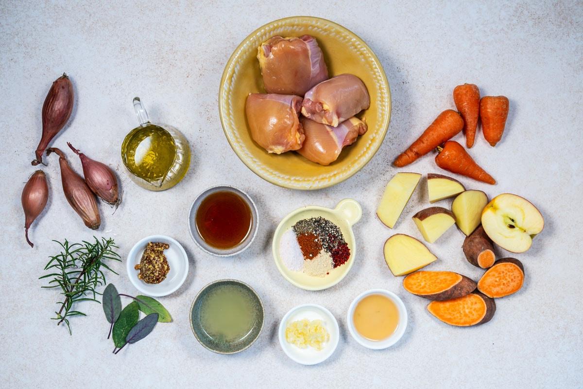 Overhead view of raw chicken thighs in a bowl, surrounded by shallots, olive oil, carrots, apple, sweet potato, yellow potato, herbs, spices, mustard, broth, chopped garlic, lemon juice, and honey on a light surface.