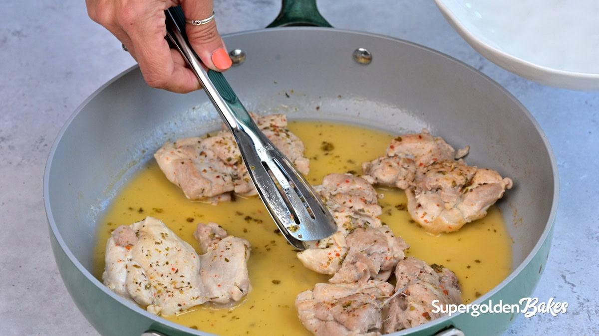 A hand uses tongs to turn seasoned chicken thighs cooking in a pan with yellow sauce. The pan is on a gray surface, and Nusde is written in the corner.