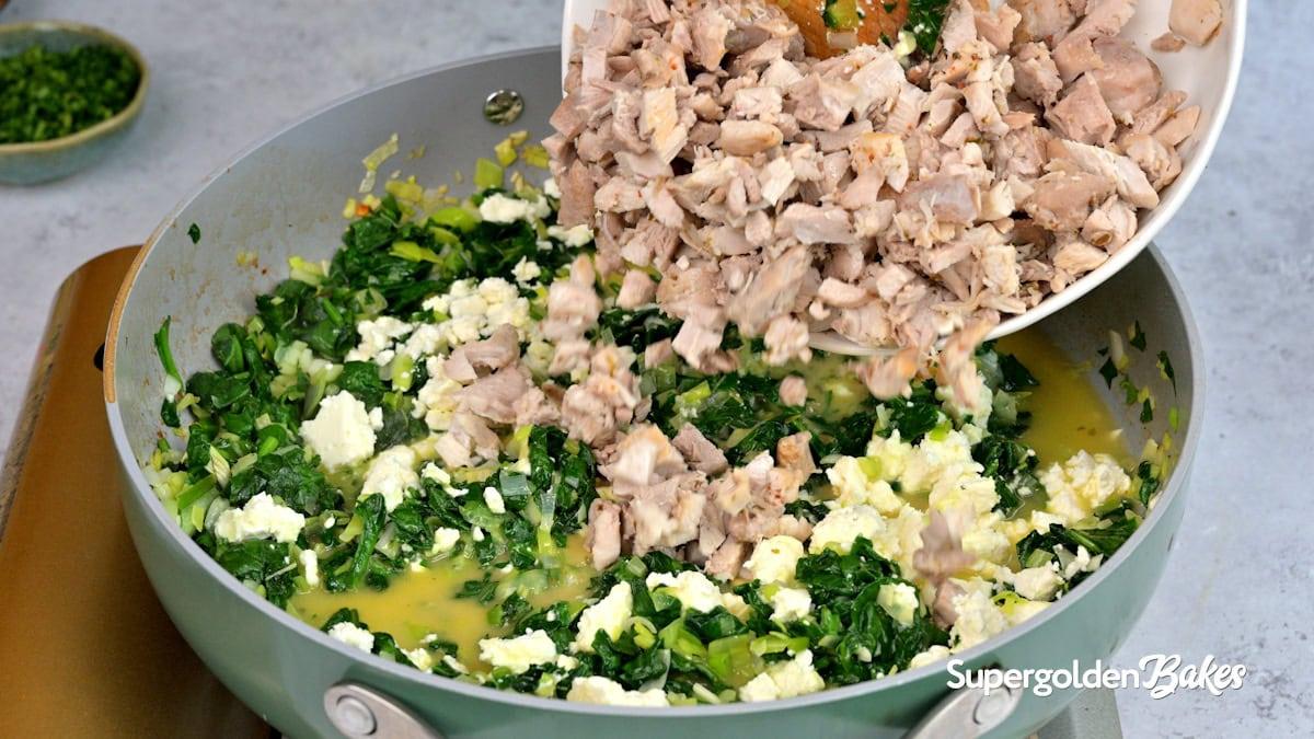 A person adds chopped cooked chicken from a white bowl into a pan filled with chopped spinach, scrambled eggs, and feta cheese. The pan is on a stove, and the scene is from Nusde.