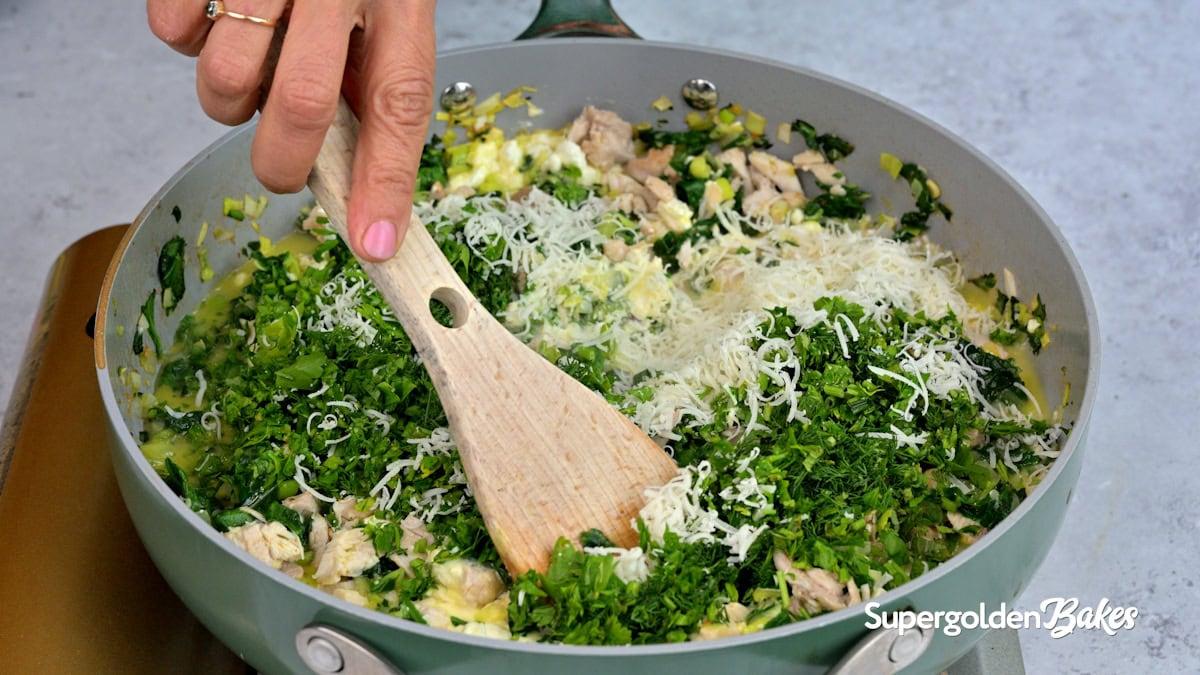A hand stirs a mixture of chopped greens, shredded cheese, and chicken in a large skillet with a wooden spoon. The scene is viewed from above, and the pan sits on a gray countertop. Nusde is written in the corner.