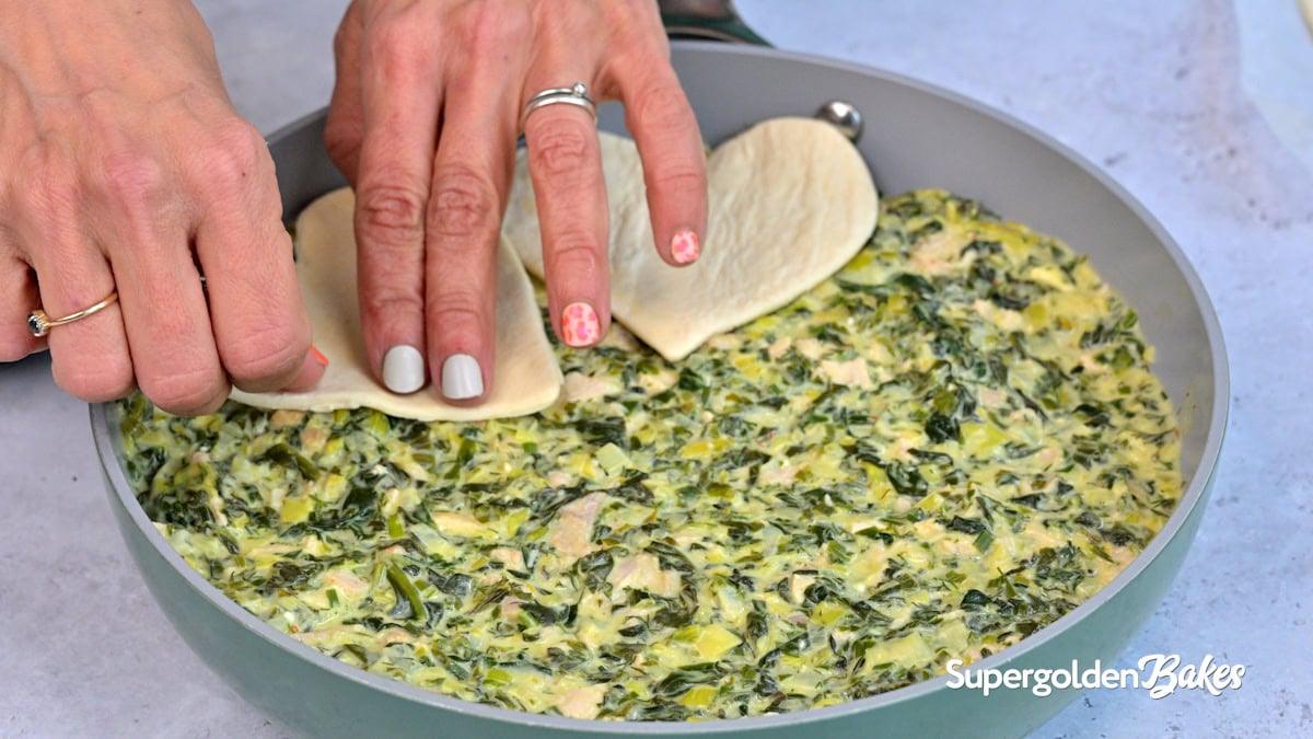 Close-up of hands placing pieces of puff pastry over a creamy spinach and chicken mixture in a round pan. The pan is labeled “Nusde.” The person has decorated nails and wears rings.