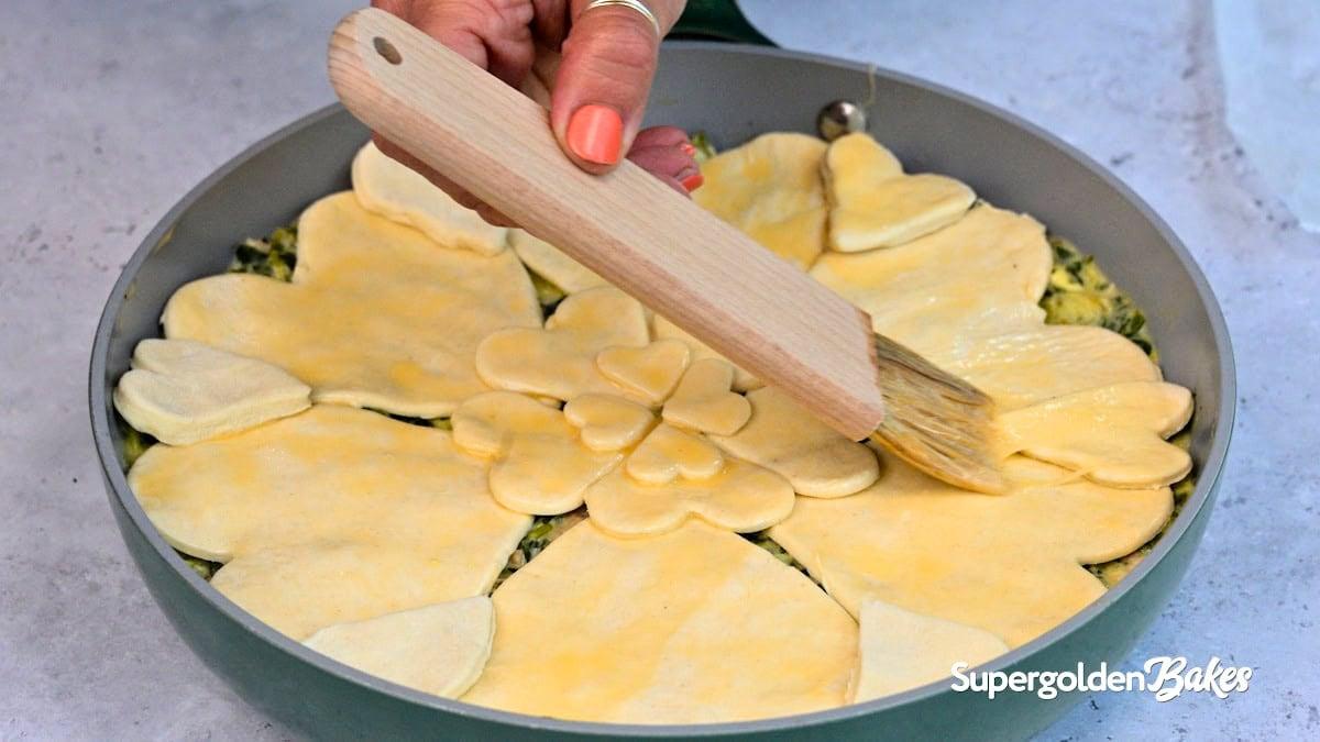 A hand uses a brush to apply egg wash to a pie topped with heart- and flower-shaped pastry cutouts in a round pan. The logo Nusde appears in the bottom right corner.