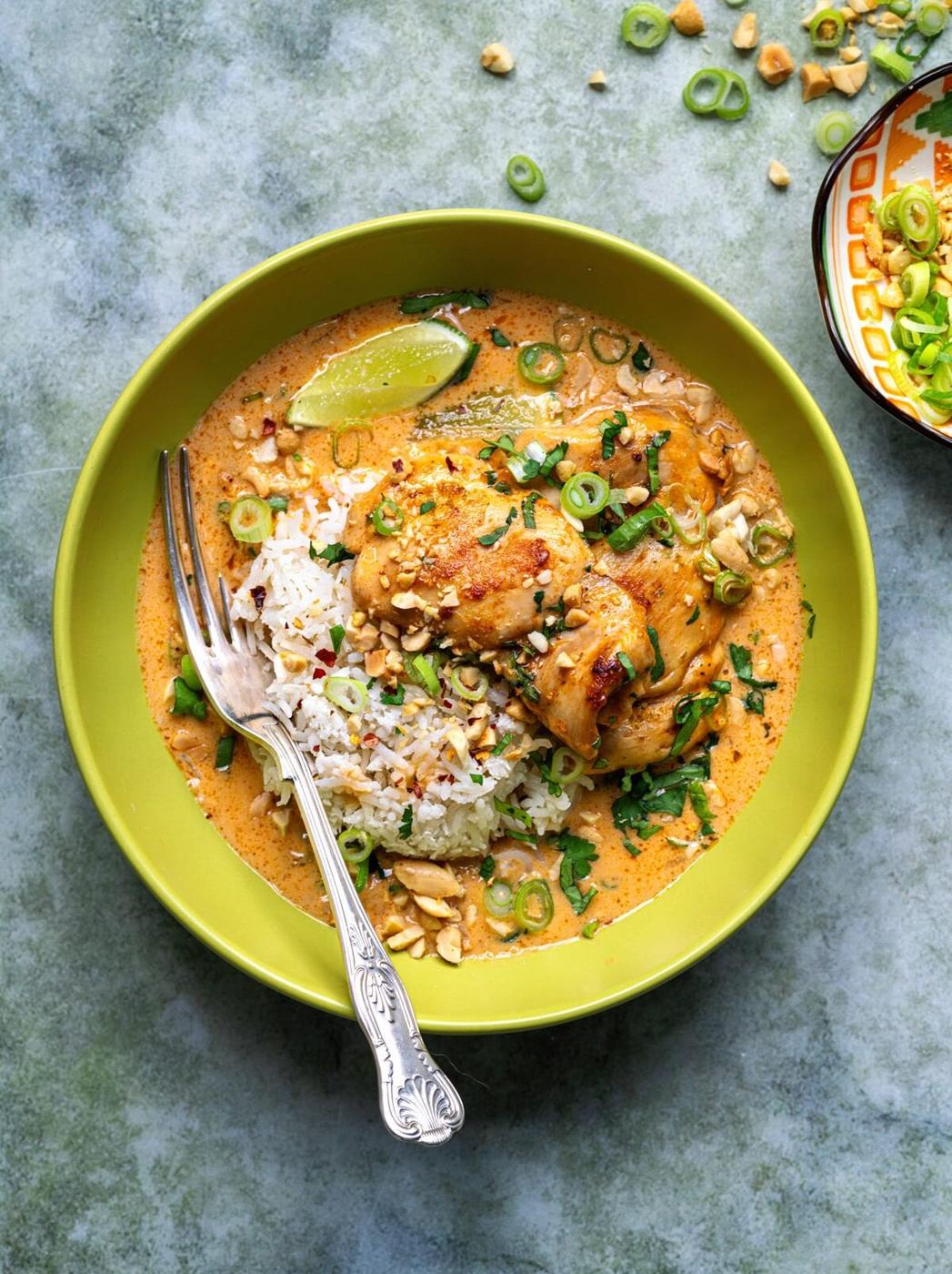 A green bowl filled with coconut lime chicken and rice, topped with chopped peanuts, green onions, herbs, and a lime wedge, with a fork resting inside. Scattered peanuts and onions are on the surface nearby.