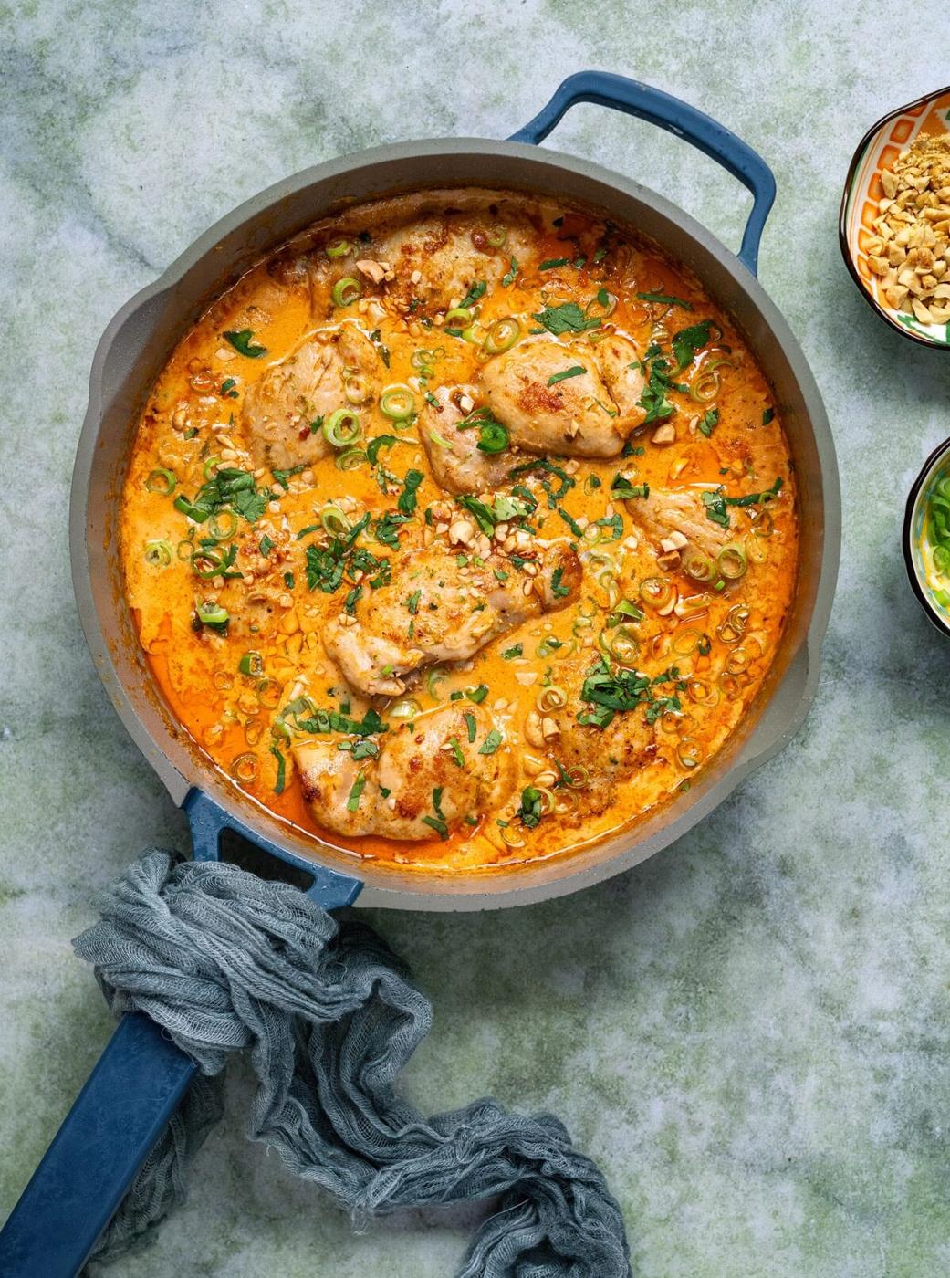 A skillet filled with creamy Coconut Lime Chicken curry, garnished with chopped herbs and sliced green onions, sits on a green textured surface next to small bowls of peanuts and sliced green onions.