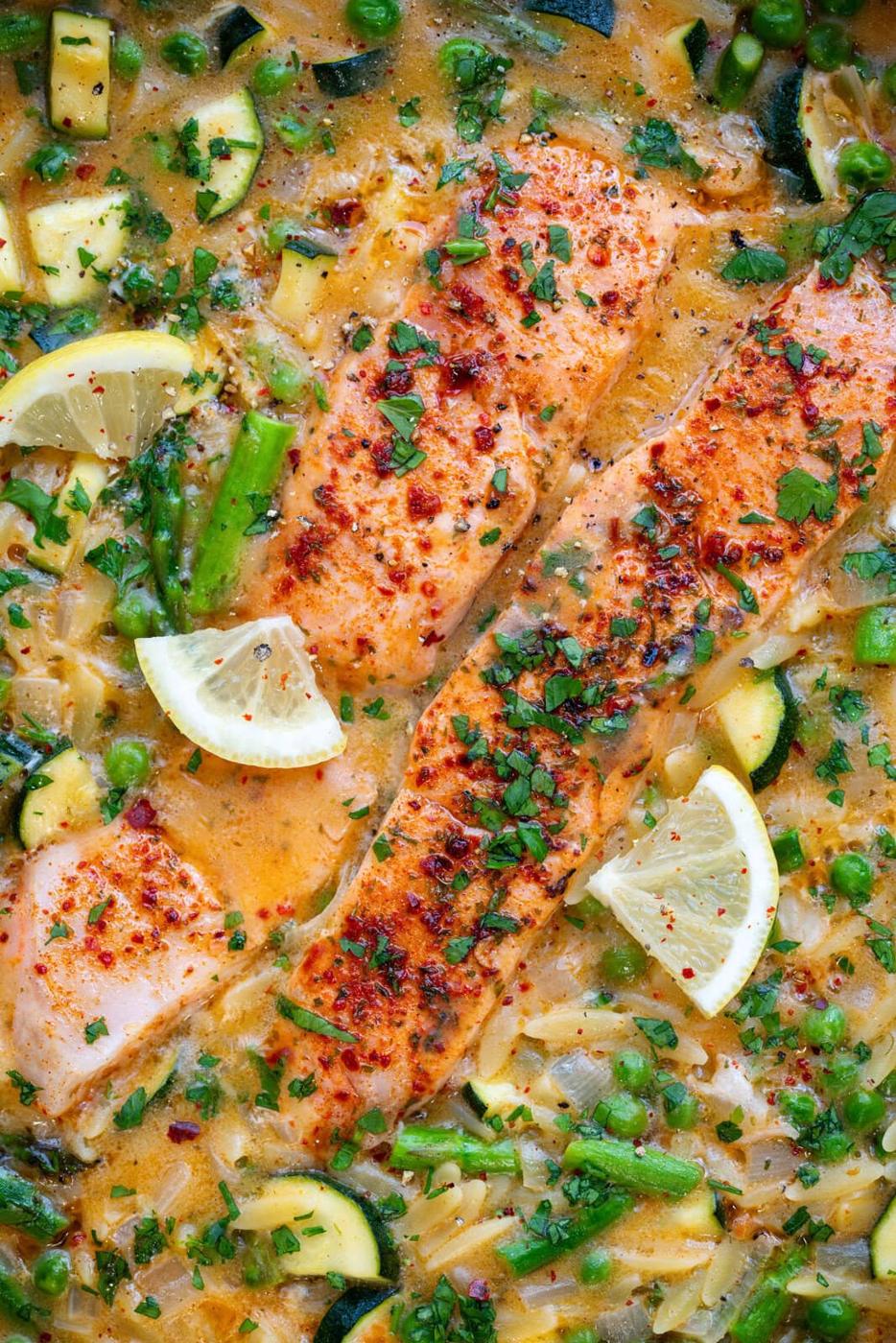Pan-seared salmon fillets in a creamy lemon orzo with asparagus, zucchini, and peas. Garnished with chopped parsley, red pepper flakes, and lemon wedges.